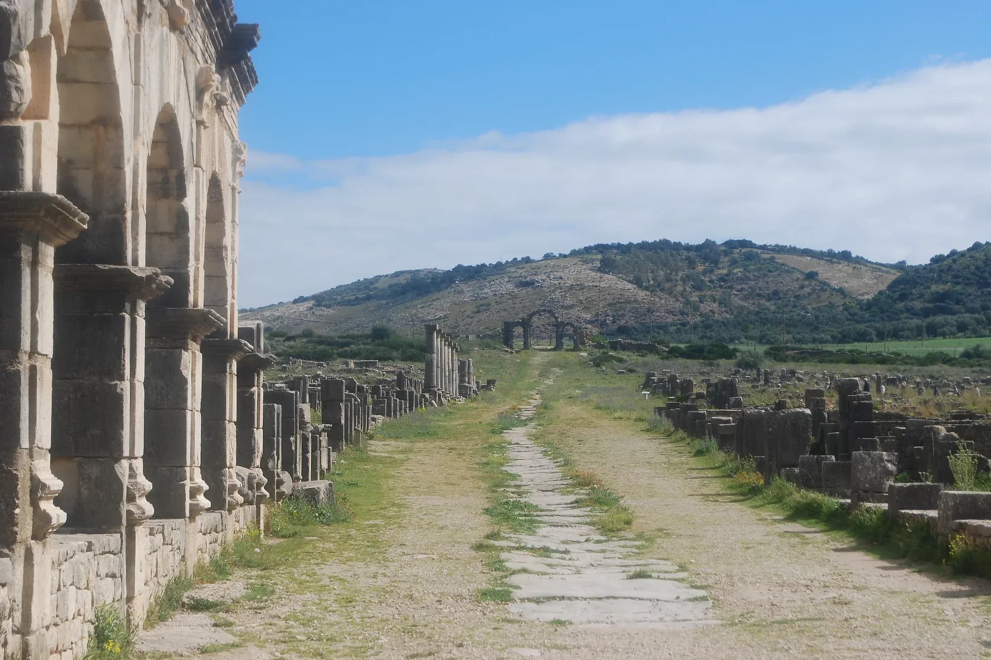 Decumano máximo de Volubilis, calle romana flanqueada por arcadas y colinas verdes