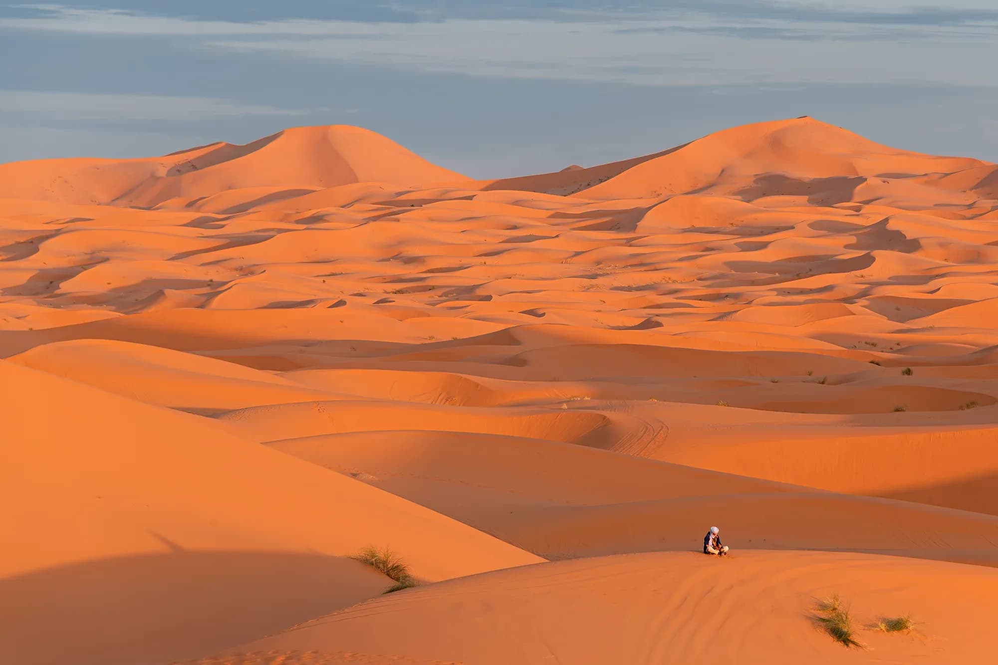 Dunas anaranjadas de Erg Chebbi con luz de tarde y una pequeña figura sentada en la arena