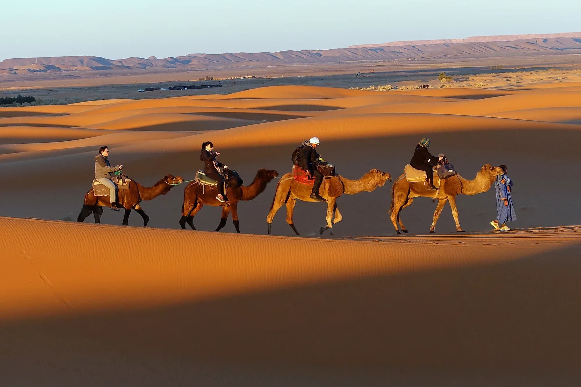 Caravana de dromedarios con viajeros cruzando las dunas de Erg Chebbi con luz cálida